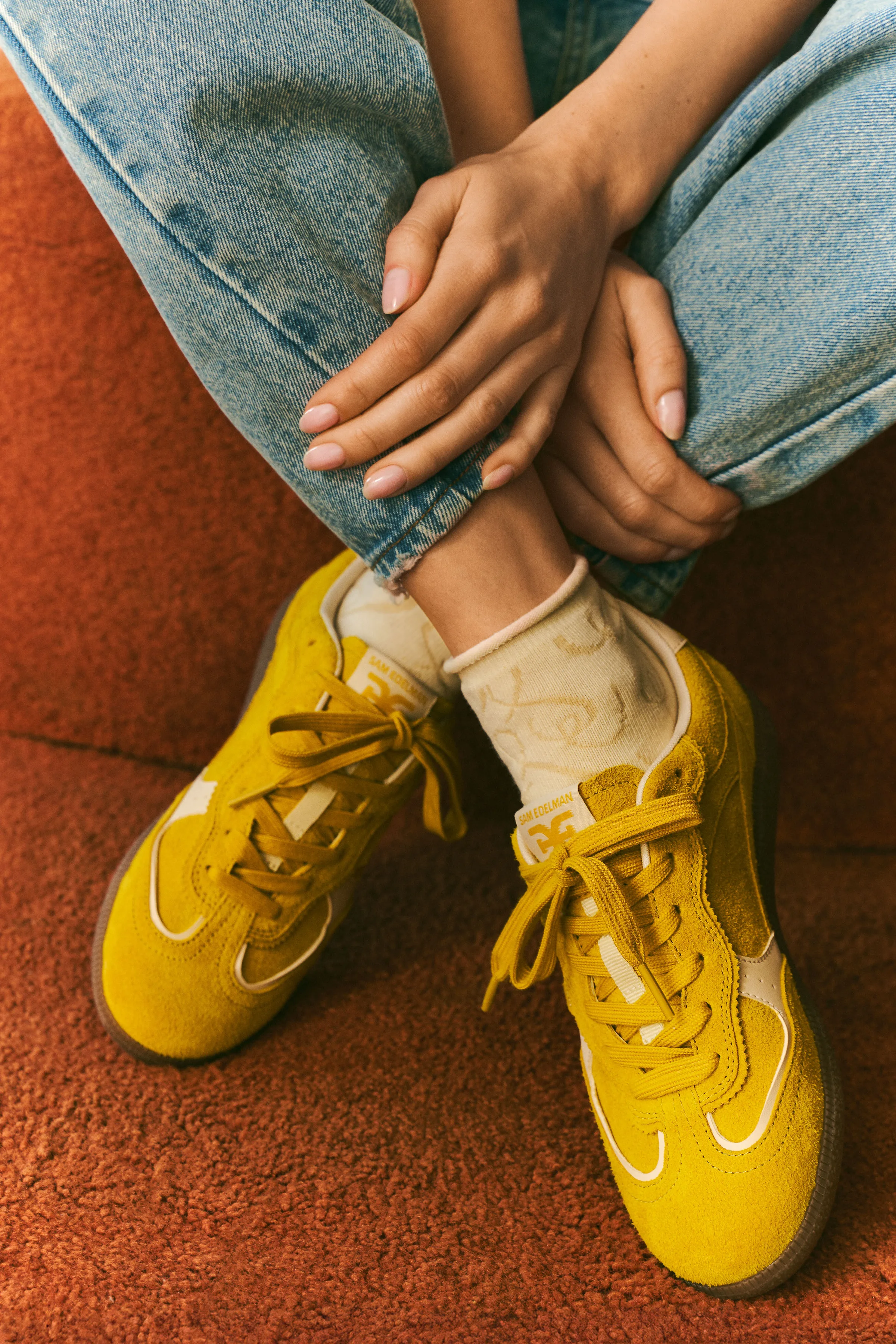 Seated model in cuffed jeans and cream socks wearing yellow suede lace-up sneakers on a rust carpet, for Sam Edelman.