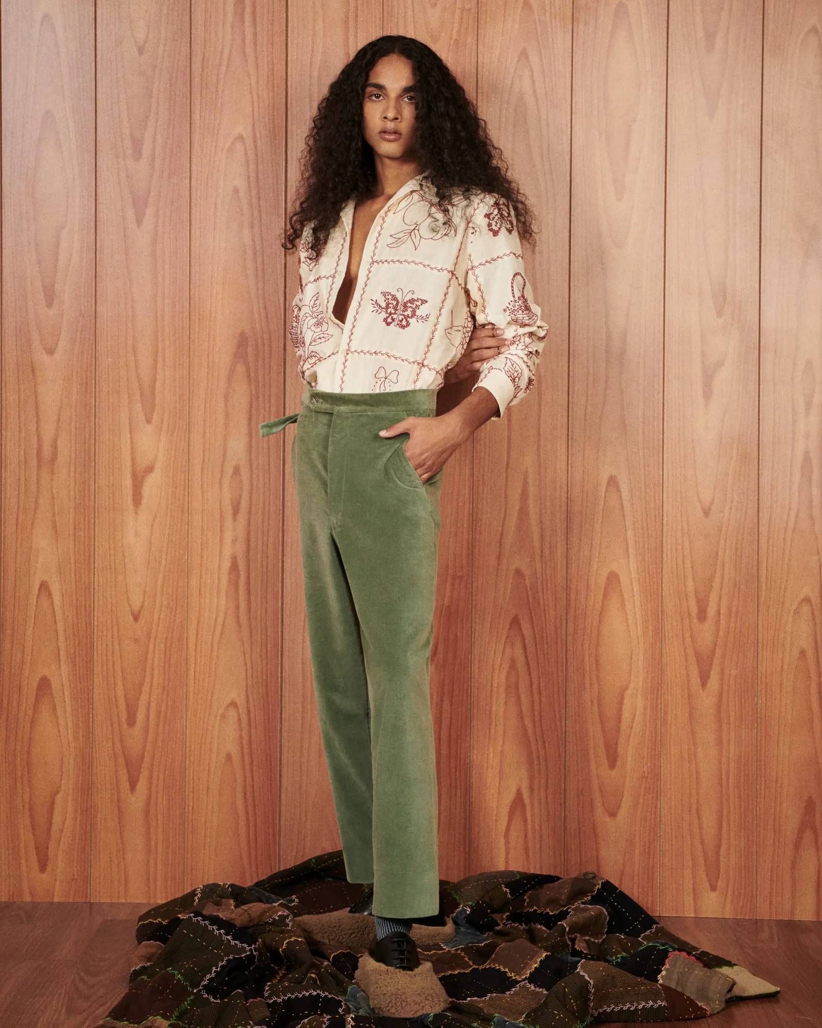 Model in embroidered cream shirt and green corduroy trousers before wood paneling, on a patchwork blanket, for The Last Magazine.