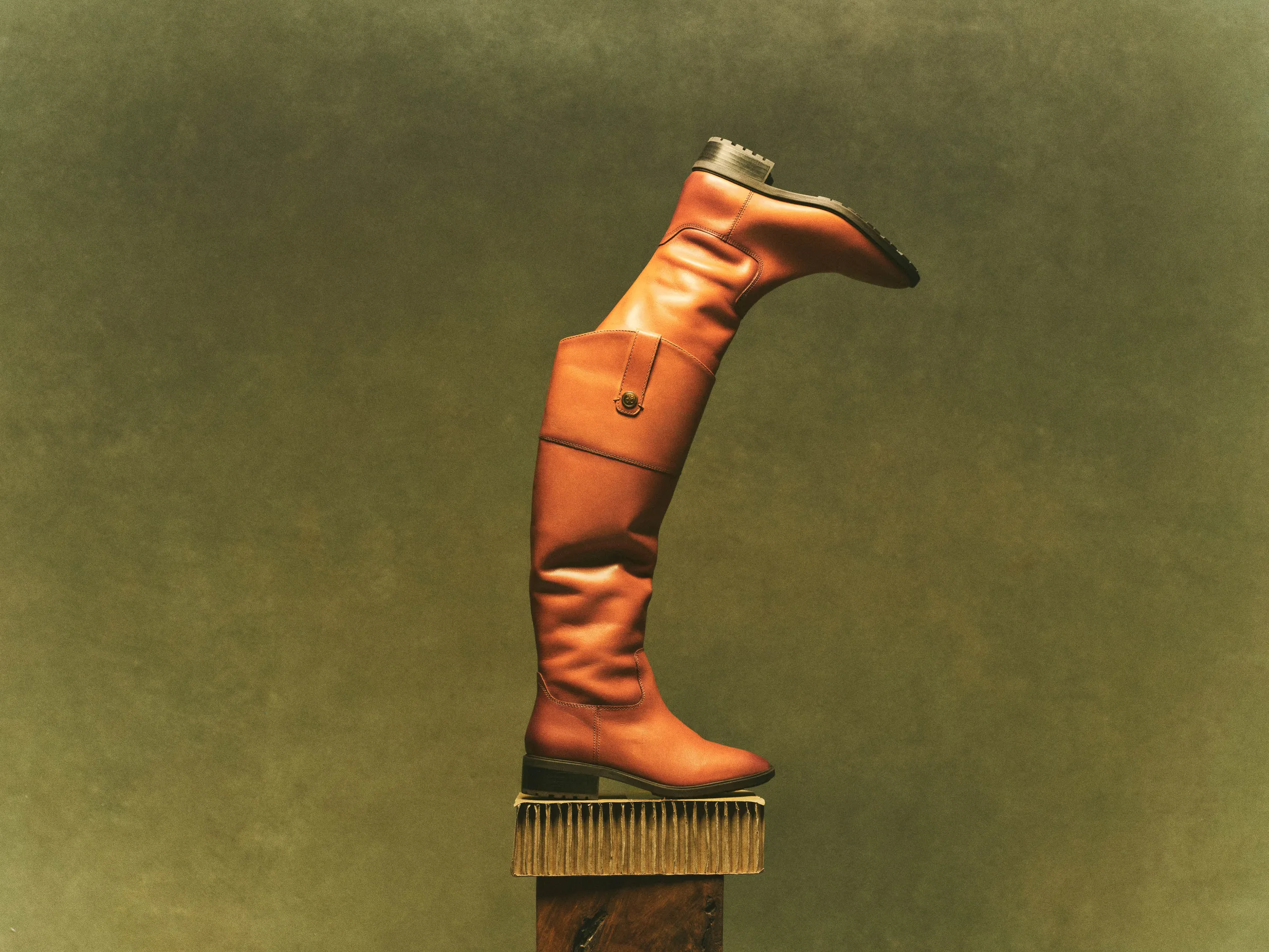 Two whiskey leather knee-high riding boots stacked on a wooden pedestal against an olive backdrop, for Sam Edelman.