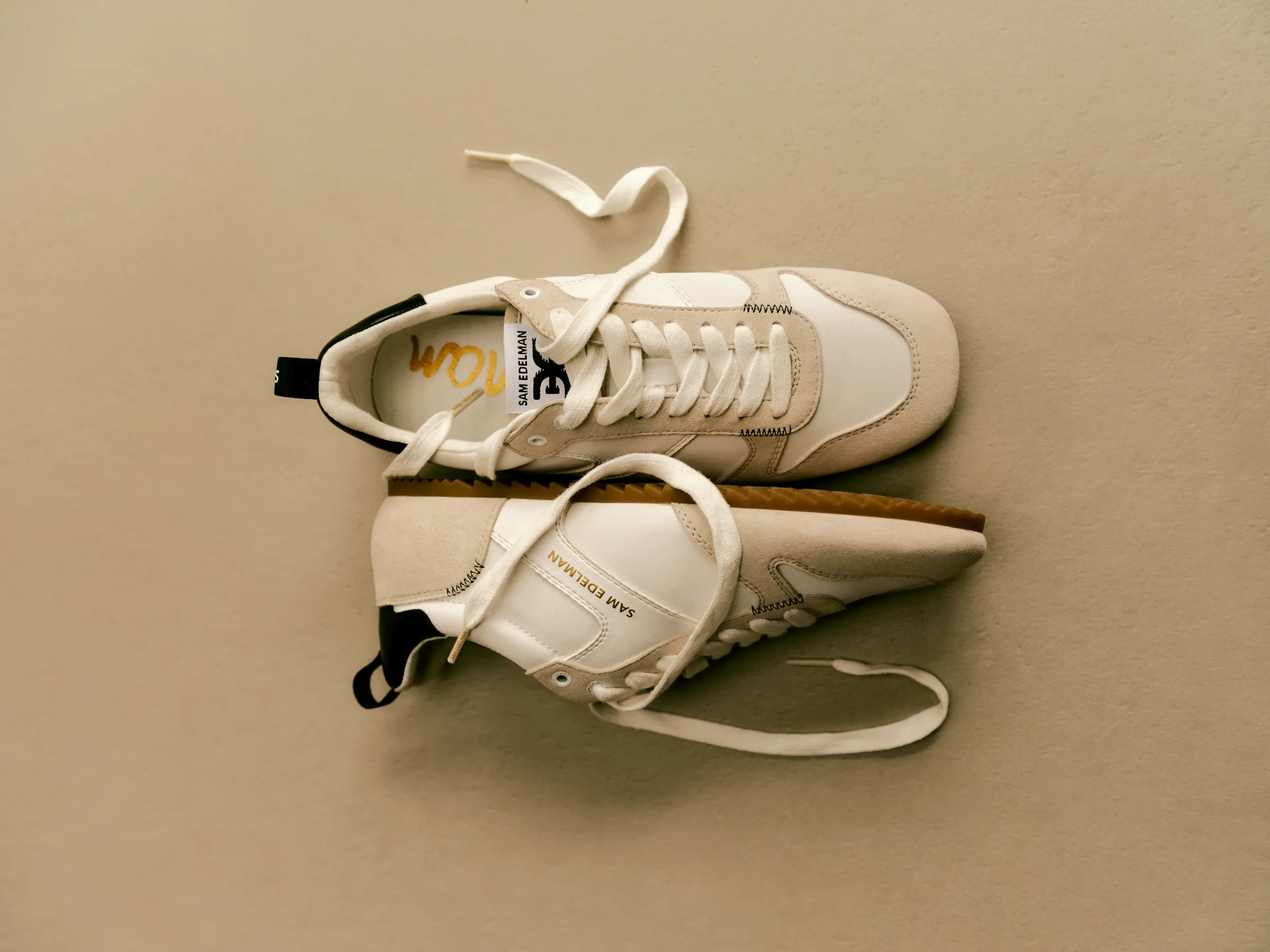 Pair of cream leather and suede retro sneakers with loose laces and gum soles on a beige backdrop, for Sam Edelman.
