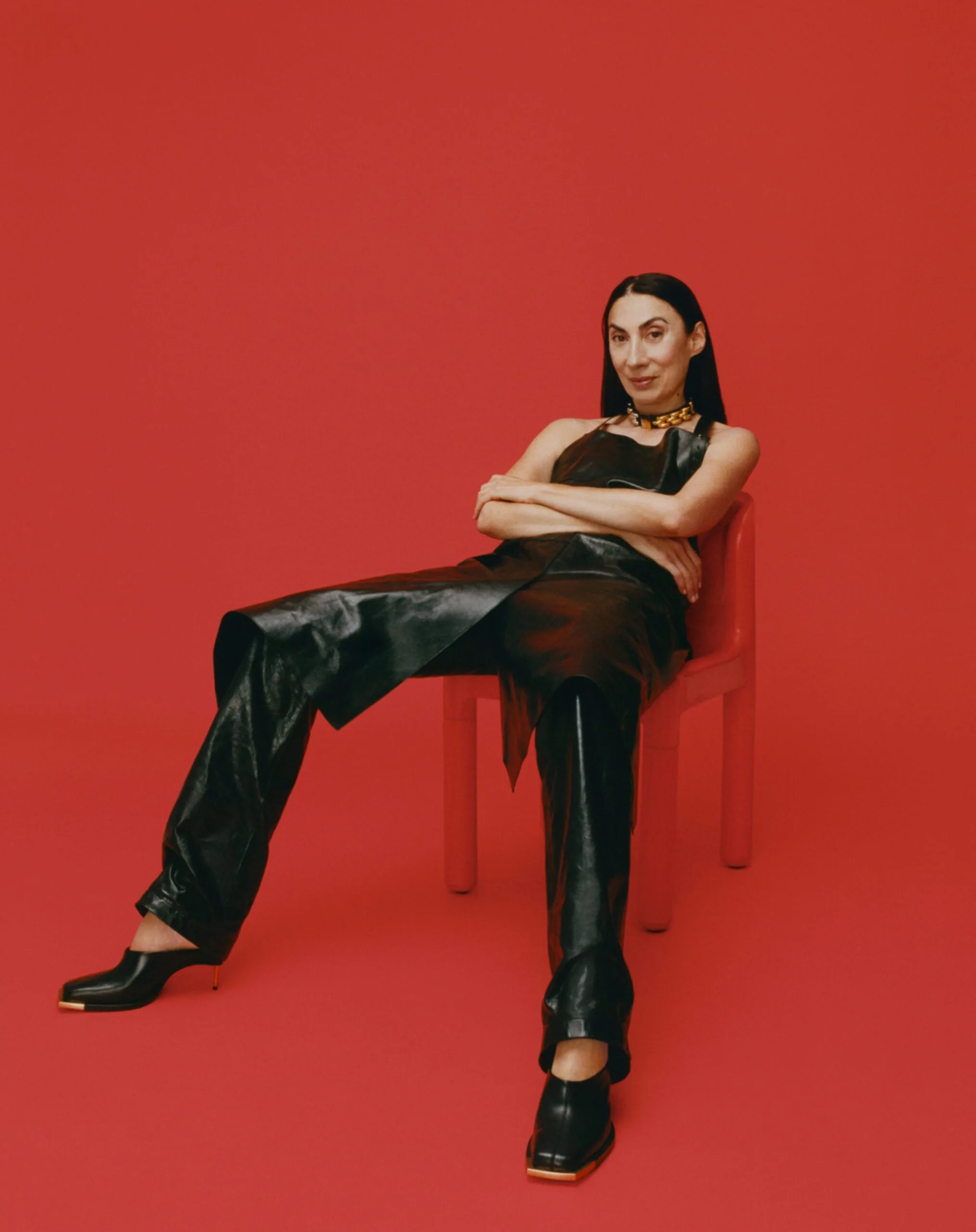 Woman in black leather apron top and trousers seated on a red chair against a red backdrop, for Interview Magazine.