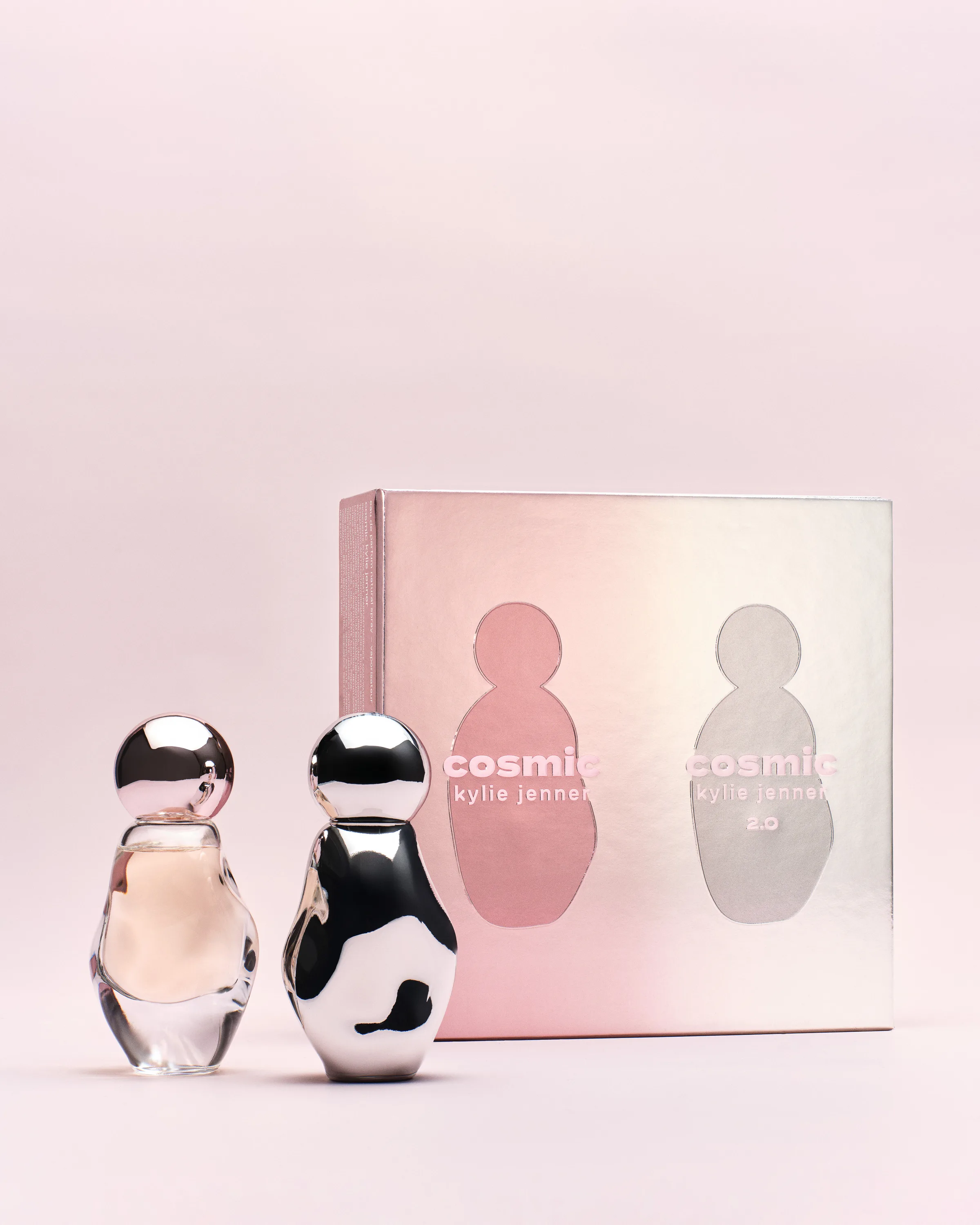 Two Cosmic perfume bottles, one clear and one chrome, beside an iridescent pink gift box on a pale pink backdrop, for Kylie Cosmetics.