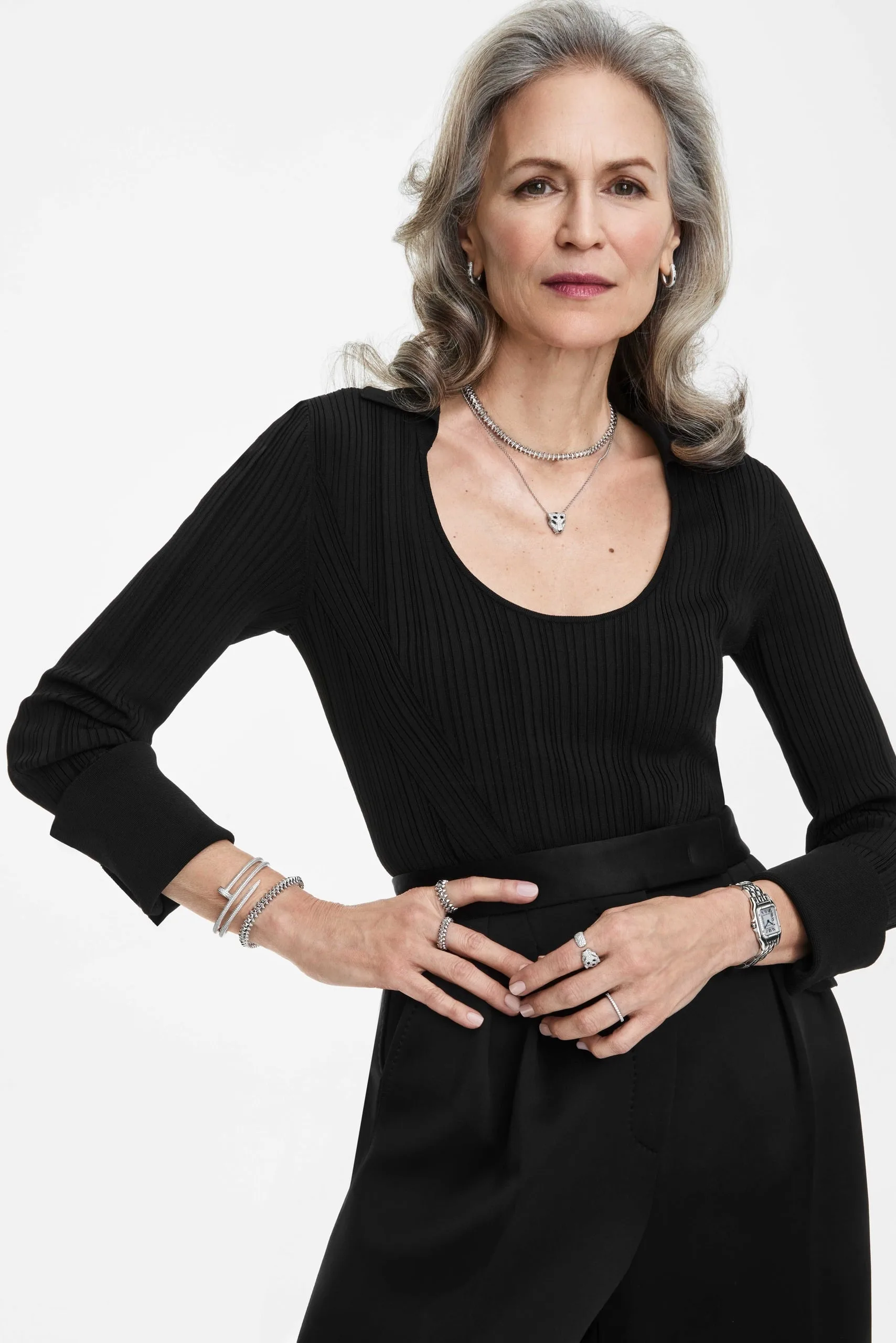 Silver-haired model in black ribbed top wearing stacked silver necklaces, bracelets, rings and watch on white, for Cartier.