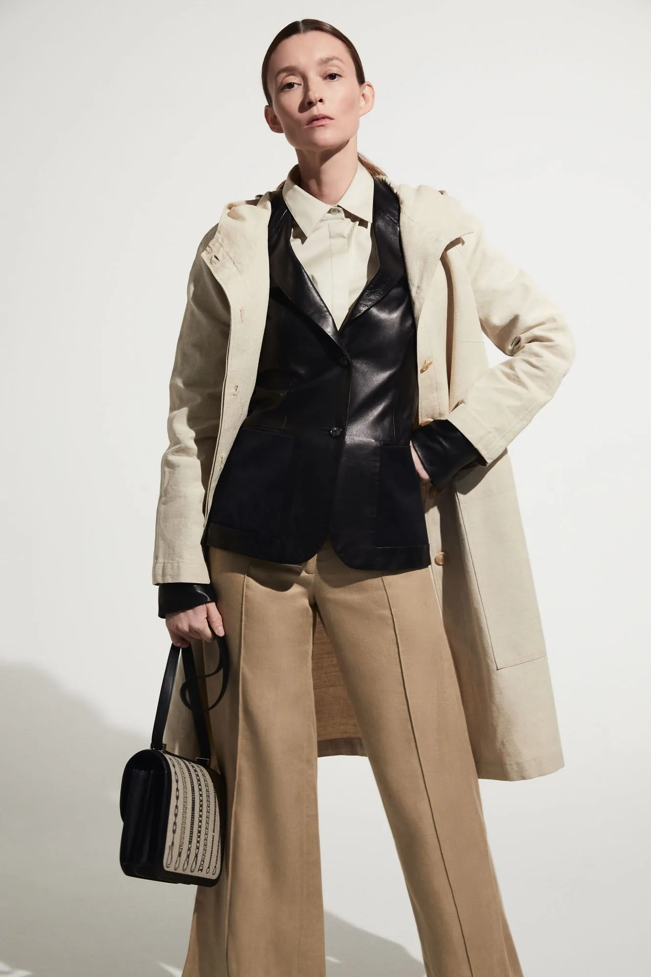 Model in a cream linen coat, black leather vest and wide camel trousers holding a woven handbag, for The RealReal.