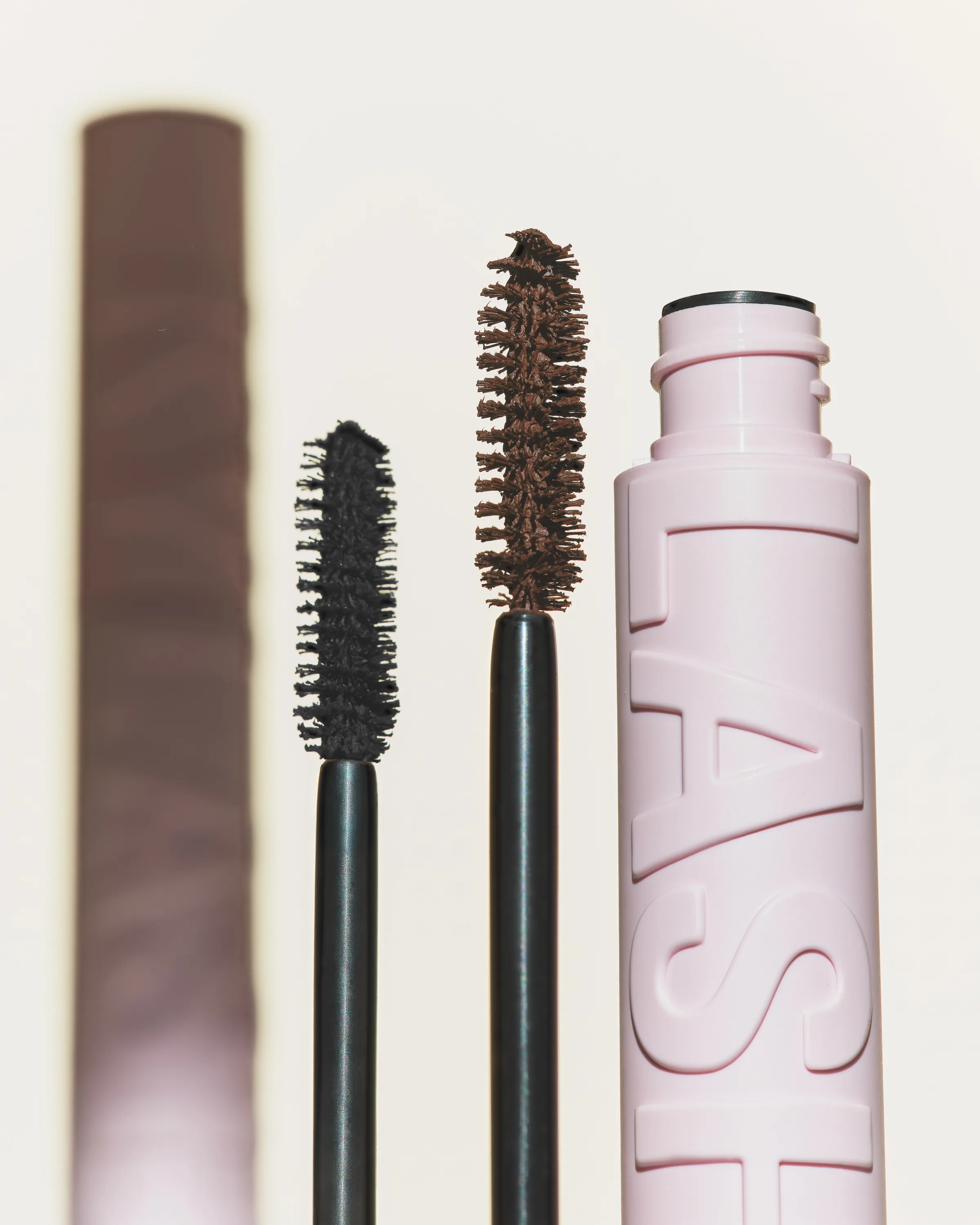 Black and brown mascara wands beside a pink embossed Lash tube on a cream background, for Kylie Cosmetics.
