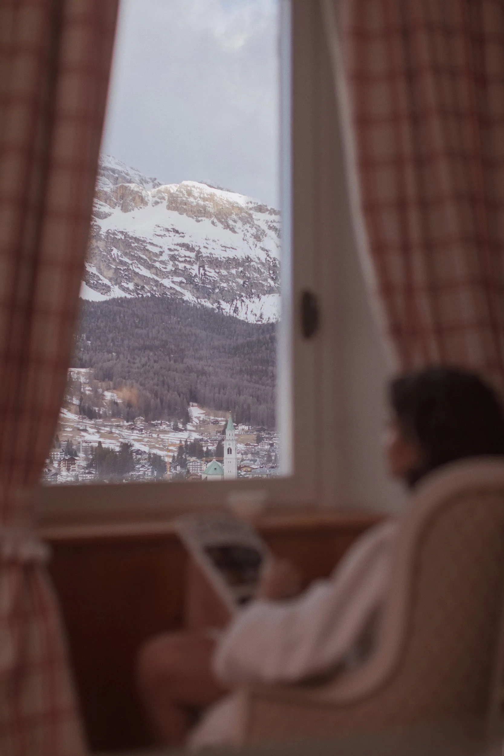 Snowy mountain and village seen through a window with plaid curtains, a blurred guest in a robe seated nearby, for Marriott.