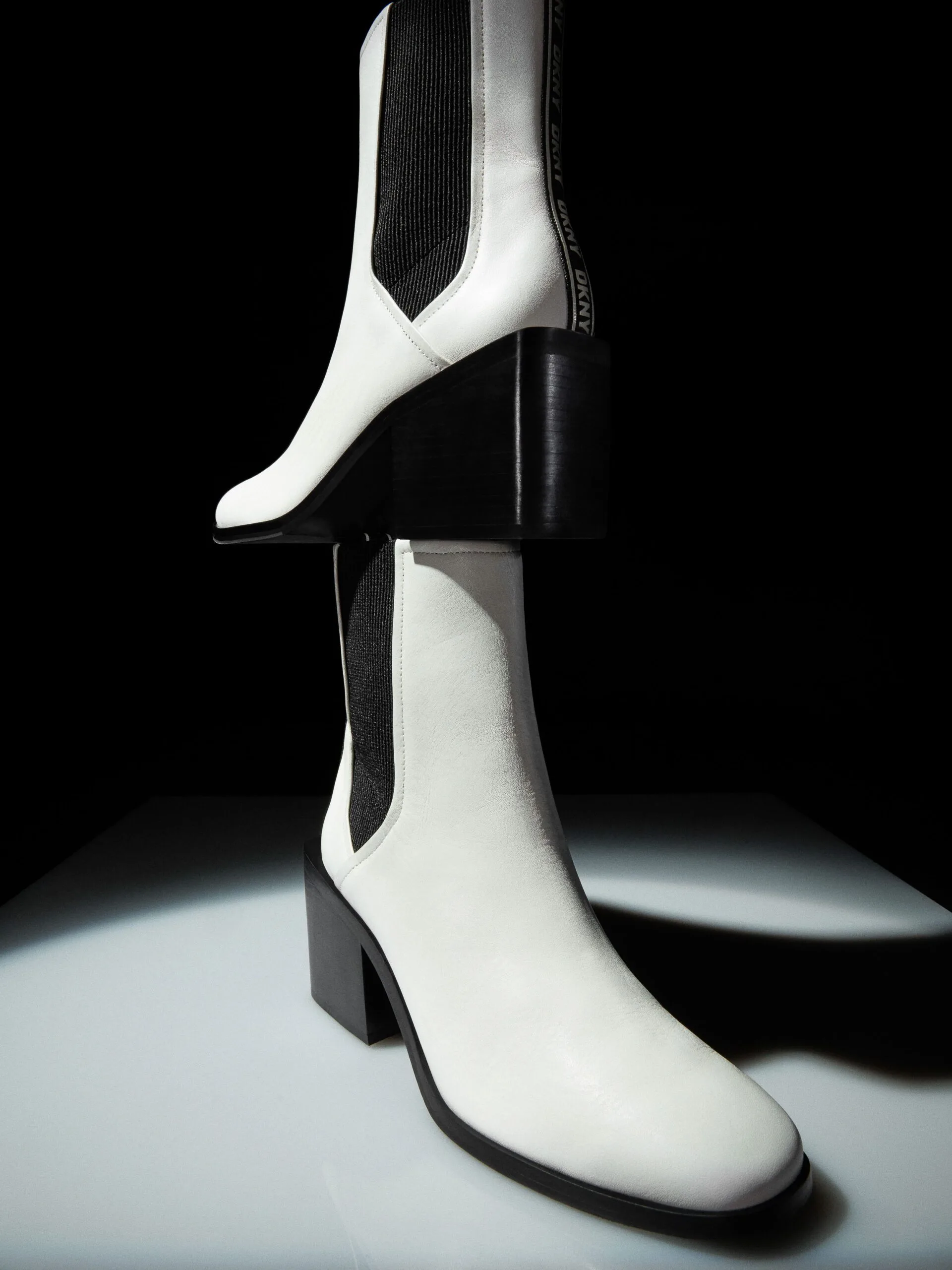 White leather Chelsea boots with black block heels stacked under a spotlight on a black backdrop, for DKNY.