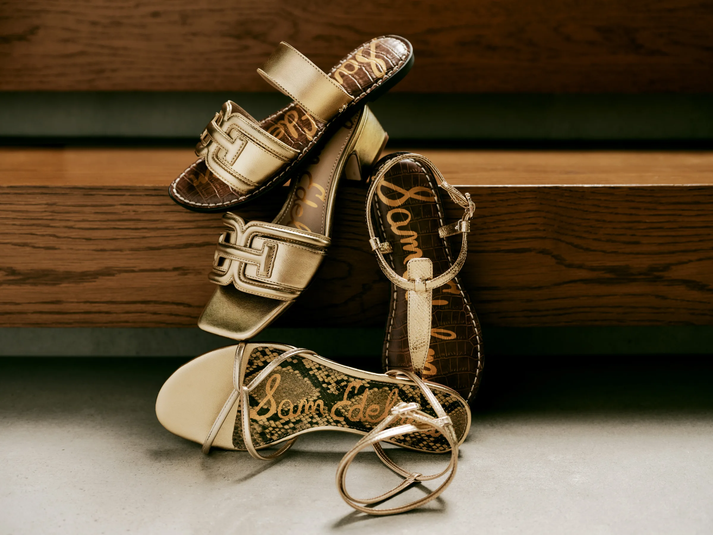 Four metallic gold sandals with logo footbeds stacked against wooden steps on a concrete floor, for Sam Edelman.