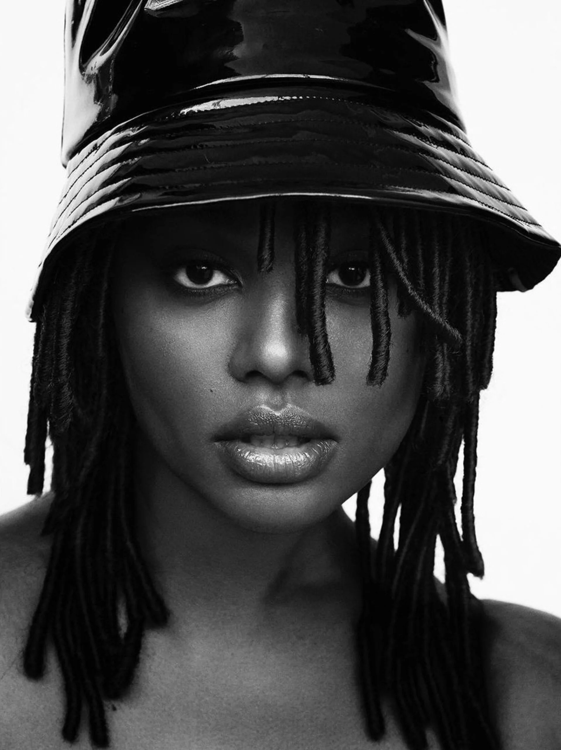Black and white portrait of a model with long locs wearing a glossy patent leather bucket hat, for V Magazine.