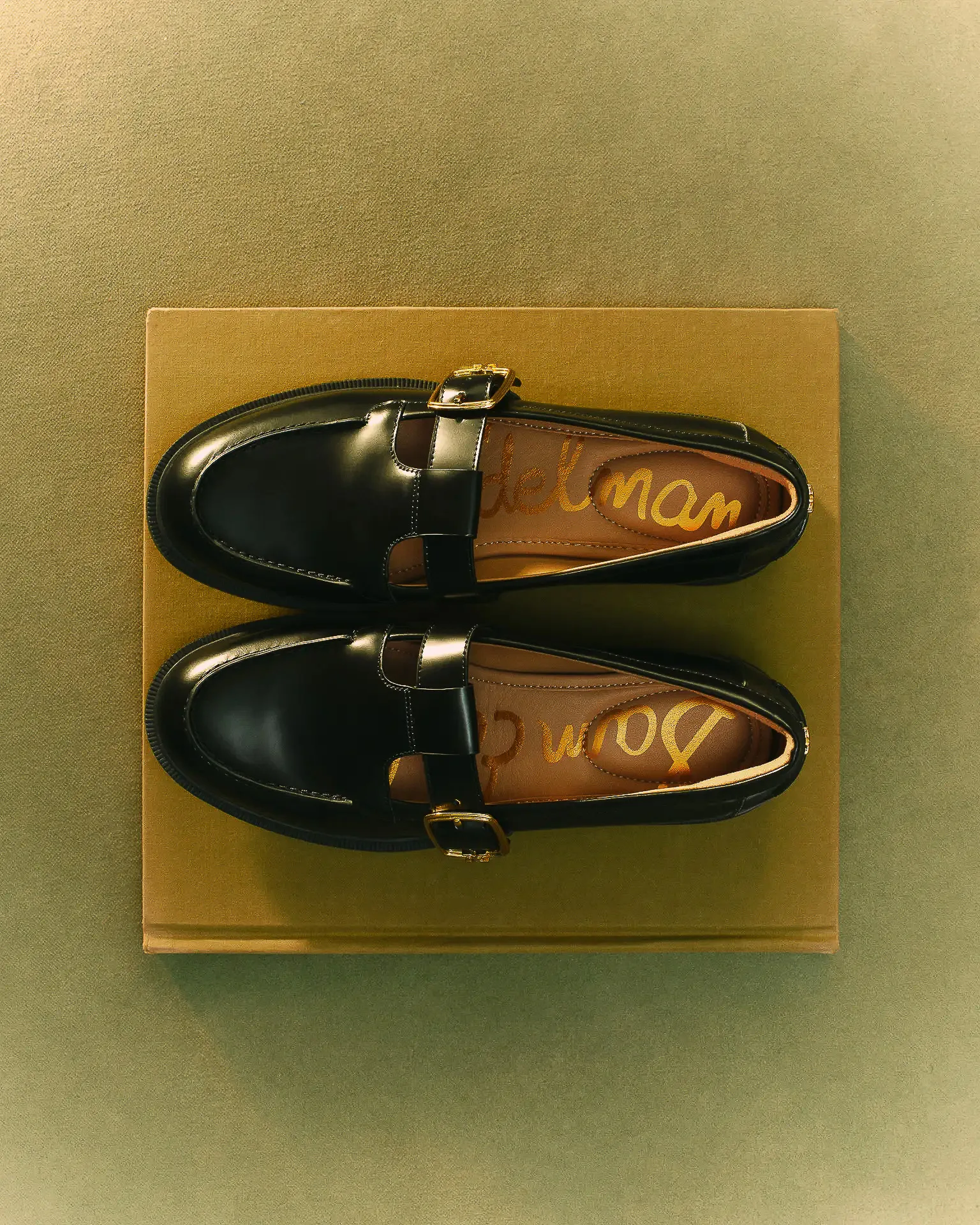 Black patent Mary Jane loafers with gold buckles on a mustard book against an olive backdrop, seen from above, for Sam Edelman.