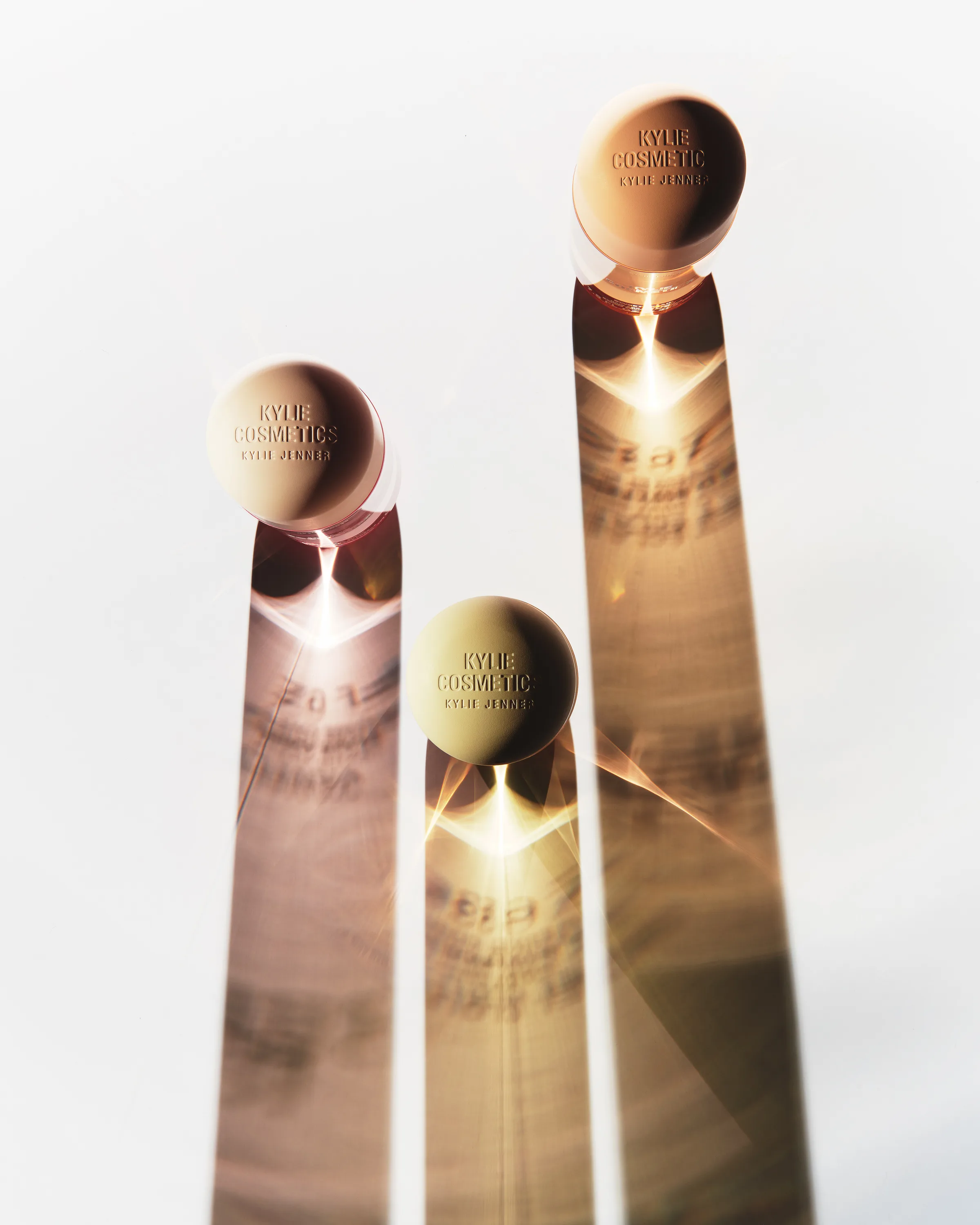 Three Kylie Cosmetics mist bottles seen from above, casting long amber shadows with light flares on white, for Kylie Cosmetics.
