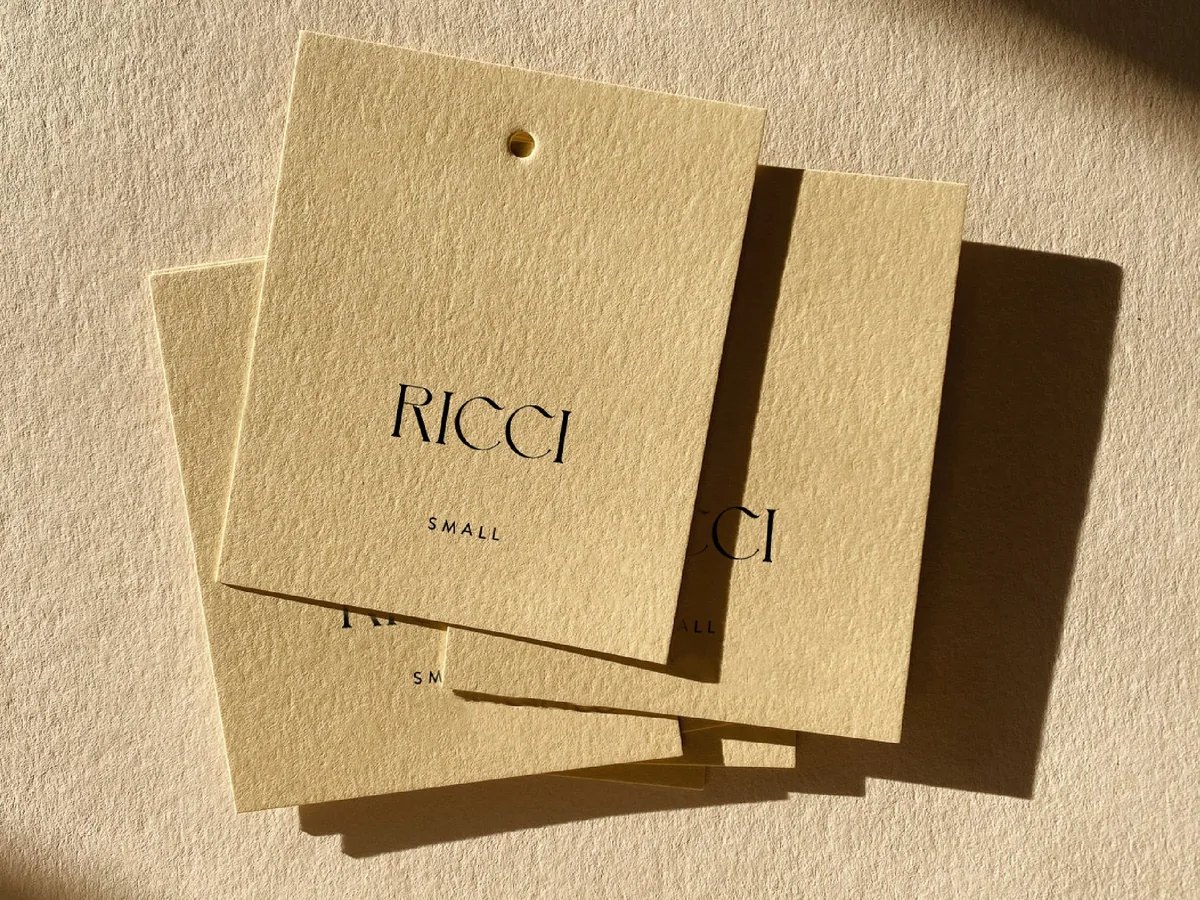 Three cream textured hang tags printed with a black serif wordmark and size Small, lying in warm sunlight, for Ricci.