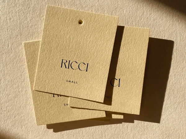 Three cream textured hang tags printed with a black serif wordmark and size Small, lying in warm sunlight, for Ricci.