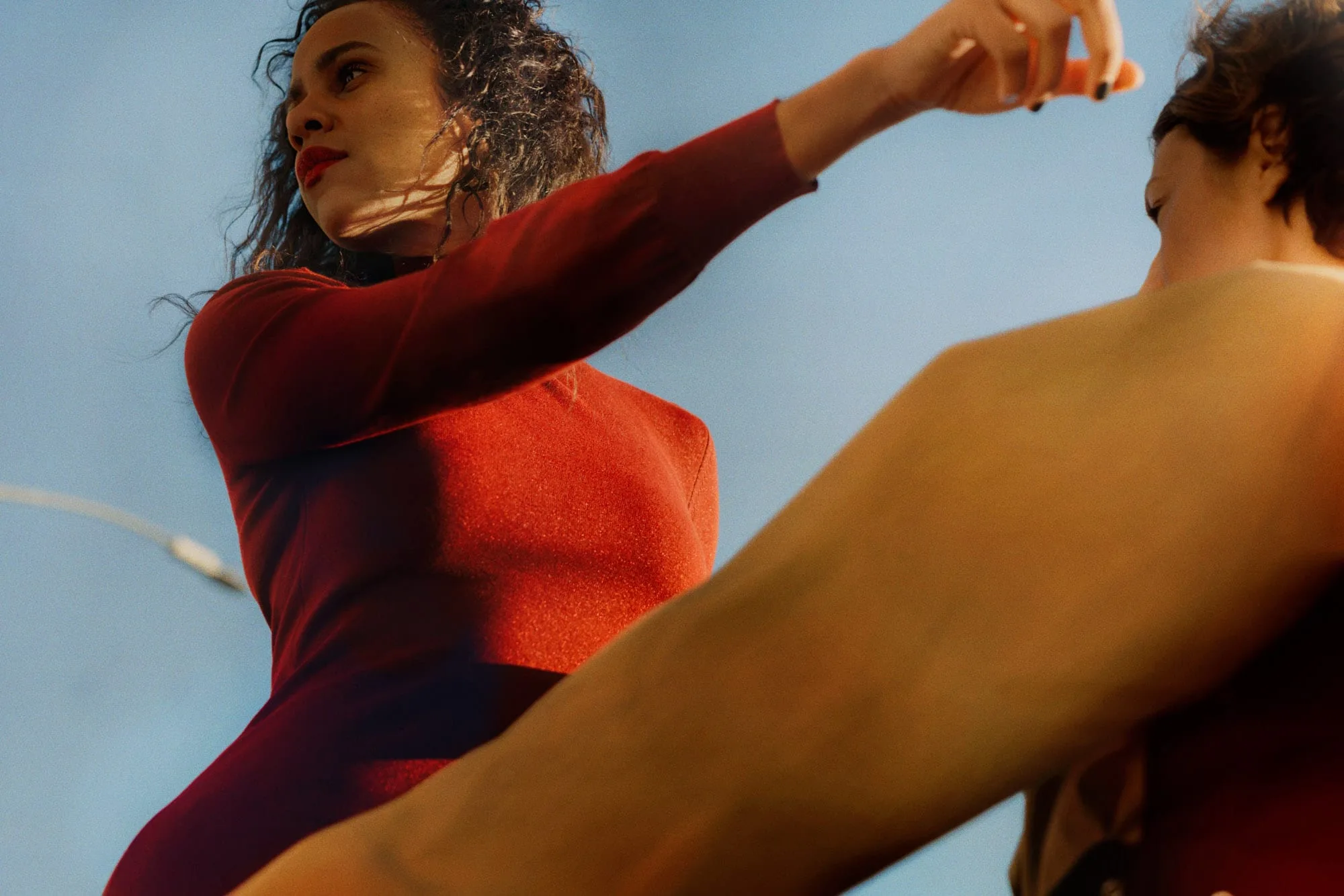 Low-angle shot of a woman in a red long-sleeve dress posing against a blue sky beside another dancer, for The Last Magazine.