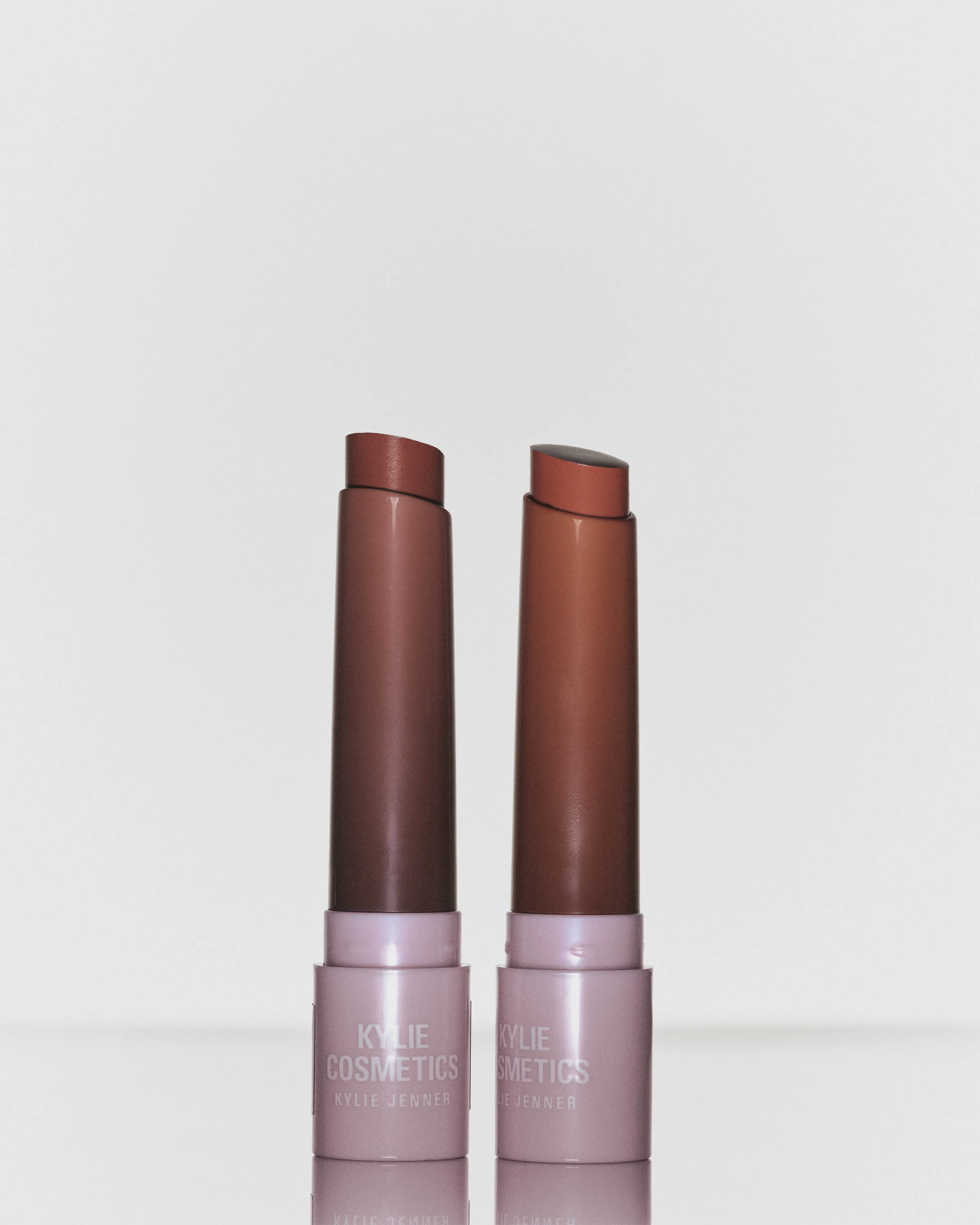 Two brown lipstick bullets in pale pink Kylie Cosmetics tubes on a light grey reflective surface, for Kylie Cosmetics.