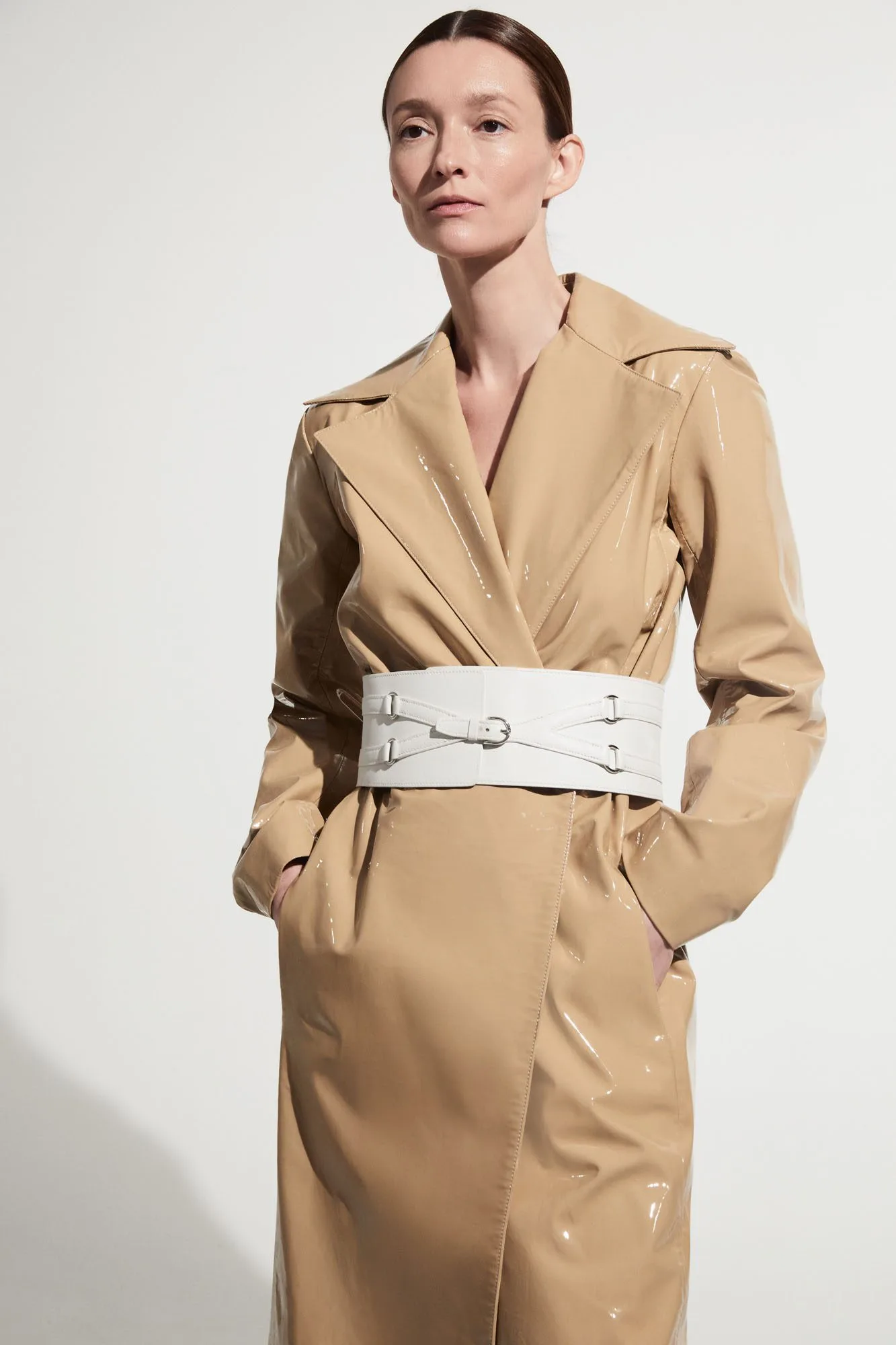 Model in a glossy beige trench coat cinched with a wide white corset belt against a white wall, for The RealReal.