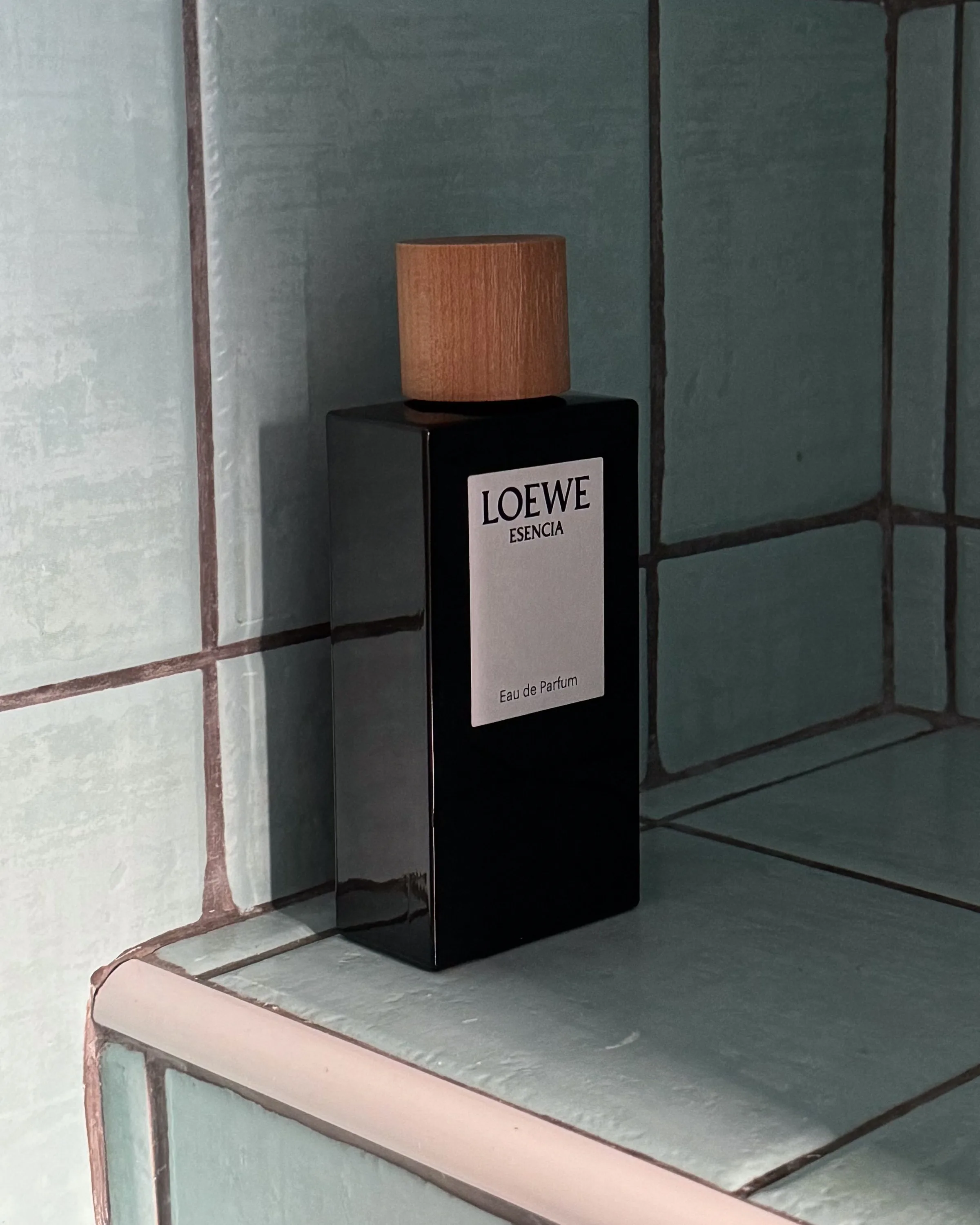 Black Loewe Esencia eau de parfum bottle with wooden cap on a pale aqua tiled bathroom ledge, for Loewe.